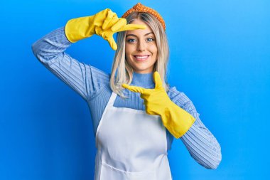 Güzel sarışın kadın daha temiz önlük ve eldivenler giyip gülümsüyor ve mutlu yüzlü eller ve parmaklarla çerçeve yapıyor. yaratıcılık ve fotoğrafçılık kavramı. 