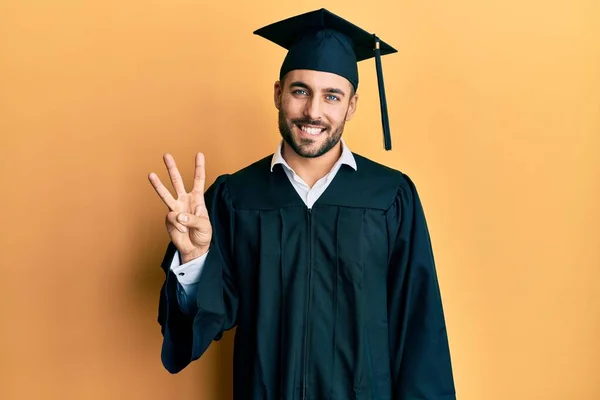 自信と幸せ笑顔ながら卒業キャップと式のローブを身に着けている若いヒスパニック系の男が現れ 指の数3で指摘します — ストック写真