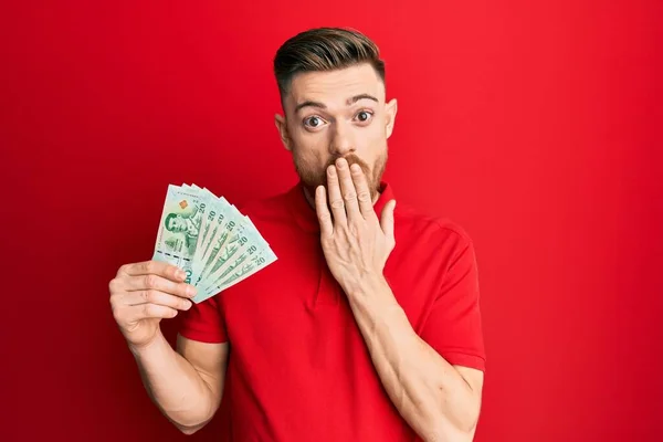 Young redhead man holding 20 thai baht banknotes covering mouth with hand, shocked and afraid for mistake. surprised expression