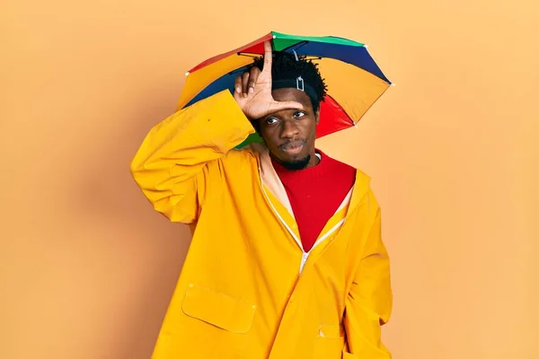 Young african american man wearing yellow raincoat making fun of people with fingers on forehead doing loser gesture mocking and insulting.