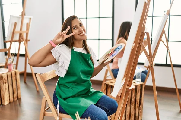 Young hispanic artist women painting on canvas at art studio doing peace symbol with fingers over face, smiling cheerful showing victory