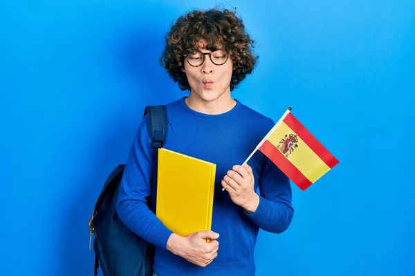 Handsome young man exchange student holding spanish flag making fish face with mouth and squinting eyes, crazy and comical.