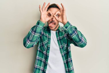 Sakallı, sıradan kıyafetler giyen, dürbün gibi hareket eden, dilini dışarı çıkaran, parmaklarından bakan yakışıklı bir adam. çılgın ifade. 