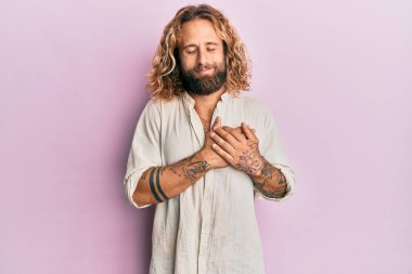 Sakallı, uzun saçlı, sıradan kıyafetler giyen, göğsünde elleri, yüzünde minnet dolu bir jestle gülümseyen yakışıklı bir adam. sağlık kavramı. 