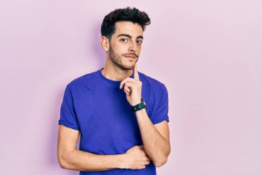 Gündelik tişört giyen genç İspanyol adam kameraya bakarken kolları çapraz ve elleri çenesinde gülümsüyor. Olumlu düşünmek. 