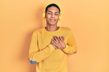Genç Afro-Amerikalı adam kulaklık takarak müzik dinliyor elleri göğsünde, gözleri kapalı gülümsüyor ve yüzünde minnet ifadesi var. sağlık kavramı. 