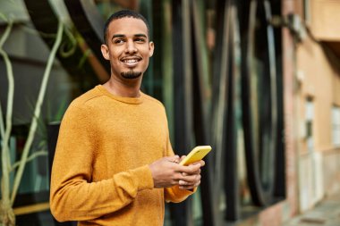 Genç Afrikalı Amerikalı adam şehirde akıllı telefon kullanarak mutlu bir şekilde gülümsüyor..