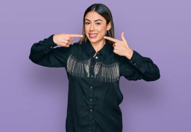 Günlük kıyafetler giyen, neşeli gülümseyen, parmaklarıyla ve ağzıyla işaret eden genç İspanyol kadın. diş sağlığı kavramı. 