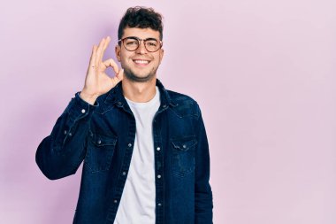 Gündelik kıyafetler ve gözlük takan genç İspanyol adam gülümsüyor. El ve parmaklarıyla 