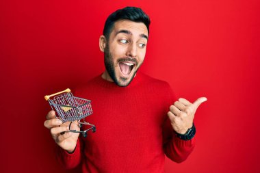 Genç İspanyol adam elinde küçük bir süpermarket arabası tutarken baş parmağını yana kaldırıyor ve ağzı açık gülümsüyor. 