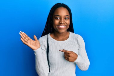 Saçları örgülü beyaz süveter giyen Afro-Amerikalı kadın kameraya hayret ediyor ve gülümsüyor.. 