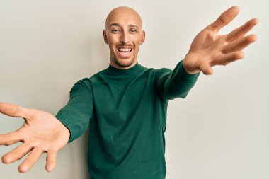 Balıkçı yaka kazak giyen kel adam kameraya sarılmak için gülümseyerek bakıyor. Mutluluğu kucaklayan neşeli bir ifade.. 