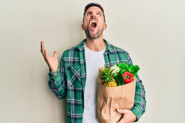 Sakallı yakışıklı bir adam elinde alışveriş torbalarıyla deli gibi bağırıyor, bağırıyor ve agresif bir ifadeyle kollarını kaldırıyor. hüsran kavramı. 