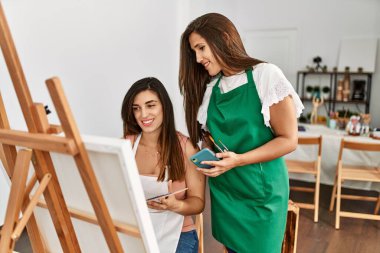 İki Latin resim öğrencisi gülümsüyor, resim yapıyor ve sanat okulunda akıllı telefon kullanıyor..