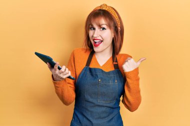 Kızıl saçlı, garson önlüğü takan, dokunmatik bir cihaz taşıyan genç bir kadın parmak ucunda, ağzı açık bir şekilde gülümsüyor. 