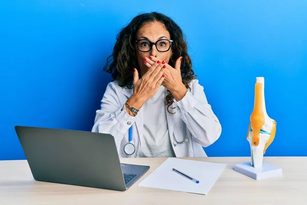 Beautiful middle age woman doctor at orthopedic clinic shocked covering mouth with hands for mistake. secret concept.