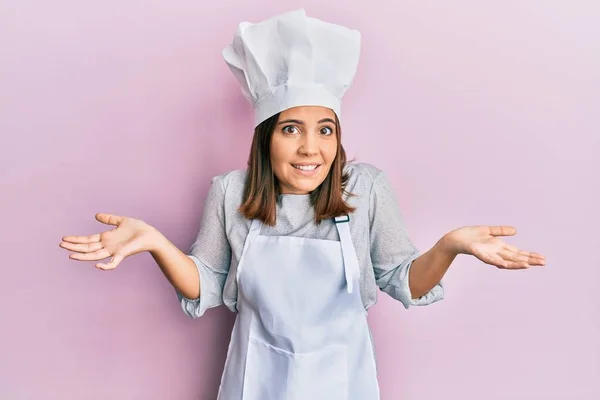 年轻美丽的女人身穿职业厨师制服 头戴无头无脑的帽子 满脸困惑 手臂和双手高举 怀疑概念 — 图库照片