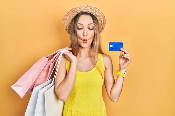 Young blonde girl holding shopping bags and credit card making fish face with mouth and squinting eyes, crazy and comical.