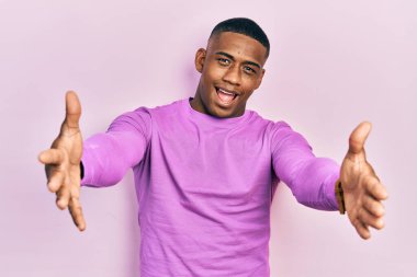 Sıradan pembe kazak giyen genç siyah bir adam kameraya bakıp sarılmak için kollarını açmış gülümsüyor. Mutluluğu kucaklayan neşeli bir ifade.. 