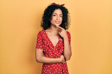 Yazlık elbise giyen genç Latin kız kameraya kendinden emin bakıyor kollarını kavuşturup gülümseyerek ve çenesini kaldırarak. Olumlu düşünmek. 