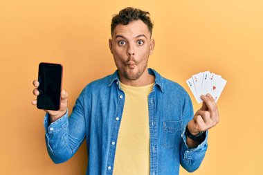Poker kartı ve akıllı telefon tutan genç İspanyol adam boş ekranı gösteriyor. Ağızlı balık suratı yapıyor ve şaşı bakıyor. Deli ve komik.. 