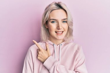 Gündelik kıyafetler giyen, neşeli gülümseyen, eli ve parmağı yana doğru olan genç sarışın kız. 