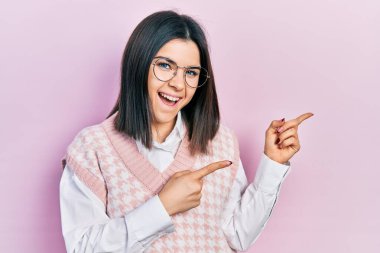 Zarif süveter giyen, gözlüklü, gülümseyen ve kameraya bakan genç esmer kadın iki elini ve parmaklarını yana doğru gösteriyor.. 
