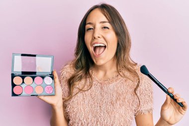 Genç esmer kadın elinde makyaj fırçası ve allık tutarak gülüyor ve kahkahalarla gülüyor.. 