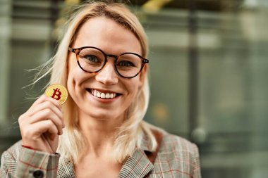 Genç sarışın iş kadını gülümseyerek şehirde Bitcoin tutuyor..