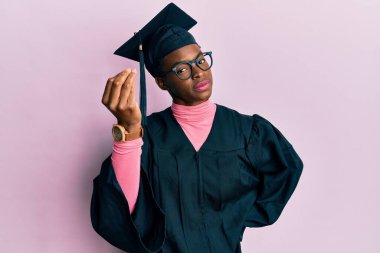 Genç Afrikalı Amerikalı kız mezuniyet şapkası ve tören cübbesi takıyor. İtalyan jesti yapıyor. El ve parmakları kendinden emin. 