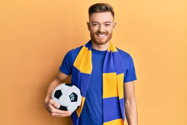 Genç kızıl saçlı futbol holiganı elinde top tutuyor. Pozitif ve mutlu görünüyor. Kendine güvenen bir gülümsemeyle dişlerini gösteriyor. 