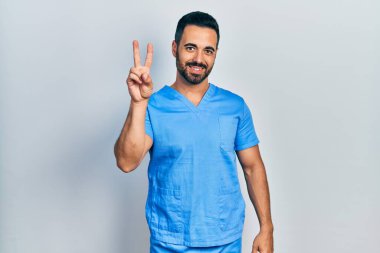 Sakallı, mavi erkek hemşire üniforması giyen yakışıklı İspanyol adam iki numaralı parmaklarıyla kendini beğenmiş ve mutlu gülümserken kendini gösteriyor.. 