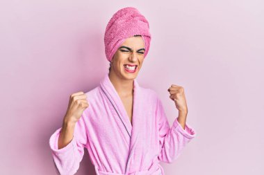 Başında duş havlusu ve bornozla makyaj yapan genç bir adam çok mutlu ve heyecanlı bir şekilde kollarını kaldırıp, gülümseyerek ve başarı çığlıkları atarak zafer jesti yapıyor. kutlama konsepti. 