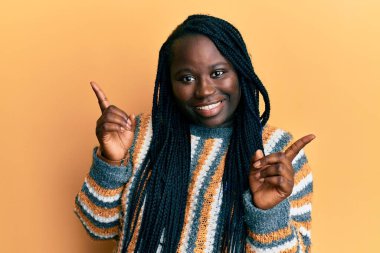 Saçları örgülü, sıradan bir süveter giyen, kendine güvenen, farklı yönleri gösteren genç siyahi bir kadın. reklam için kopyalama alanı 