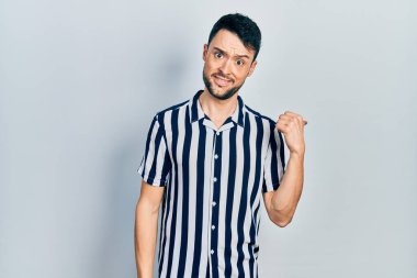 Gündelik kıyafetler giyen, gülümseyen, gülümseyen ve başparmağıyla yan tarafı gösteren İspanyol genç adam.. 