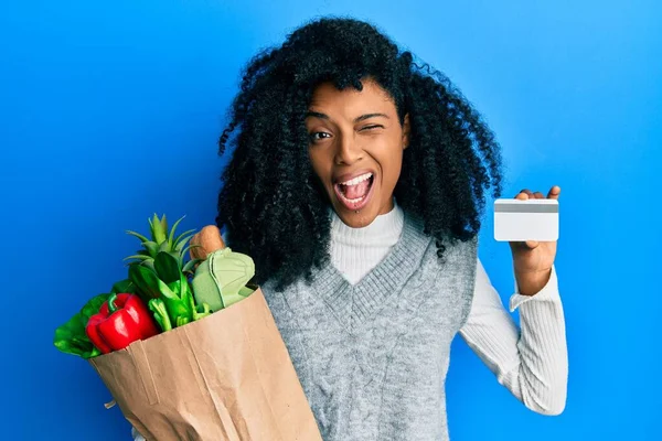 アフリカ系アメリカ人女性とともにアフロヘア保持食料品やクレジットカードのウィンク見ますカメラとともにセクシー表現 陽気で幸せな顔 — ストック写真