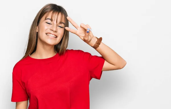 ティーンCaucasian女の子身に着けていますカジュアル赤Tシャツ行います平和シンボルとともに指上の顔 笑顔陽気に示す勝利 — ストック写真