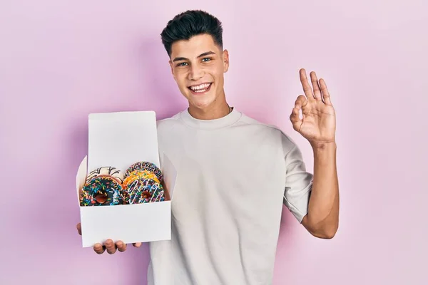 Joven Hombre Hispano Sosteniendo Caja Sabrosas Rosquillas Coloridas Haciendo Signo —  Fotos de Stock