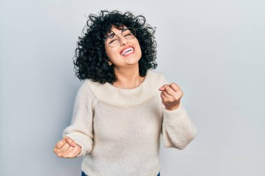 Orta Doğulu genç bir kadın, gündelik beyaz tişört giyiyor çok mutlu ve heyecanlı, kazanan bir jest yapıyor, kolları havada, gülümsüyor ve başarı için bağırıyor. kutlama konsepti. 