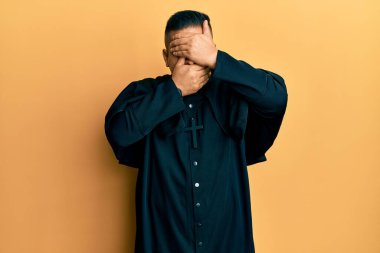 Sarı arka planda dikilen genç latin rahip gözlerini ve ağzını ellerle kapadı, şaşırdı ve şok oldu. duygu gizleme 