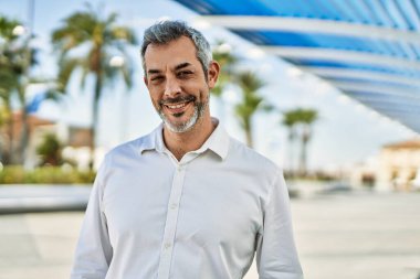 Orta yaşlı, kır saçlı, gülümseyen bir adam şehirde mutlu bir şekilde duruyor..