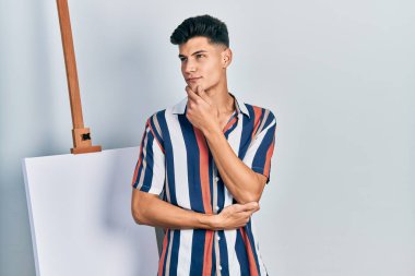 Elleri çenesinde boş tuvalin yanında durup soru soran, düşünceli bir genç adam. Düşünceli bir yüzle gülümsüyor. şüphe kavramı. 
