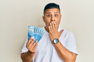 On bin Şili pezosu tutan genç latin adam ağızlarını eliyle kapatıyor, şaşırmış ve hata yapmaktan korkuyor. şaşırmış ifade 