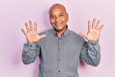 Orta yaşlı, gündelik kıyafetler giyen Latin bir adam on numarayı gösterip işaret ederken kendinden emin ve mutlu bir şekilde gülümsüyor.. 