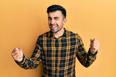 Gündelik kıyafetler giyen genç İspanyol adam çok mutlu ve heyecanlı. Kazanan jestler yapıyor. Kolları havada, gülümsüyor ve başarı için bağırıyor. kutlama konsepti. 