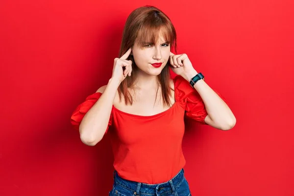 红头发的年轻女人穿着宽松的红色T恤 用手指捂住耳朵 对吵闹的音乐表示恼怒的表情 聋人概念 — 图库照片