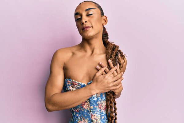 Hispanic man wearing make up and long hair wearing elegant corset smiling with hands on chest with closed eyes and grateful gesture on face. health concept.