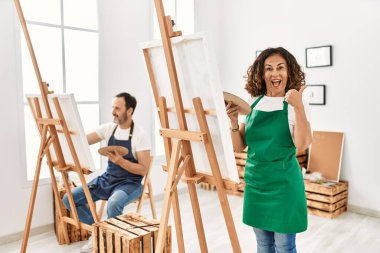 İspanyol orta yaşlı kadın ve sanat stüdyosunda olgun bir adam baş parmağını yana kaldırmış, ağzı açık gülümsüyor. 