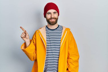 Sakallı, sarı yağmurluk giyen, yüzünde kocaman bir gülümseme olan beyaz bir adam, kameraya el ile işaret ediyor.. 