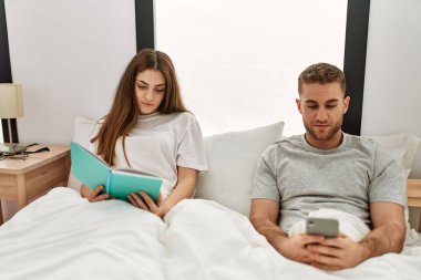 Genç beyaz çift kitap okuyor ve yatakta akıllı telefon kullanıyor..